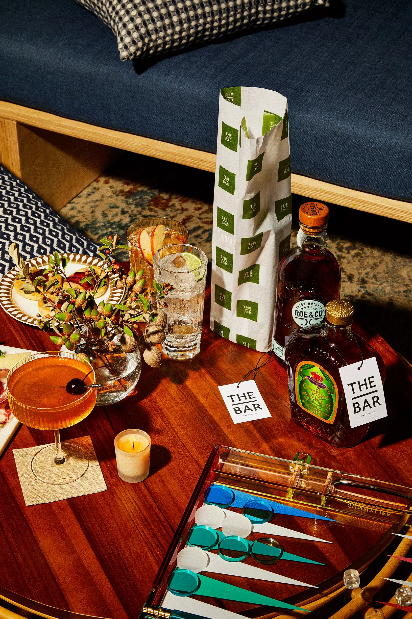 A wooden table with a cocktail, a glass of water, a plant, a candle, bottles of liquor, two The Bar coasters, and a backgammon board, set in a cozy lounge setting.