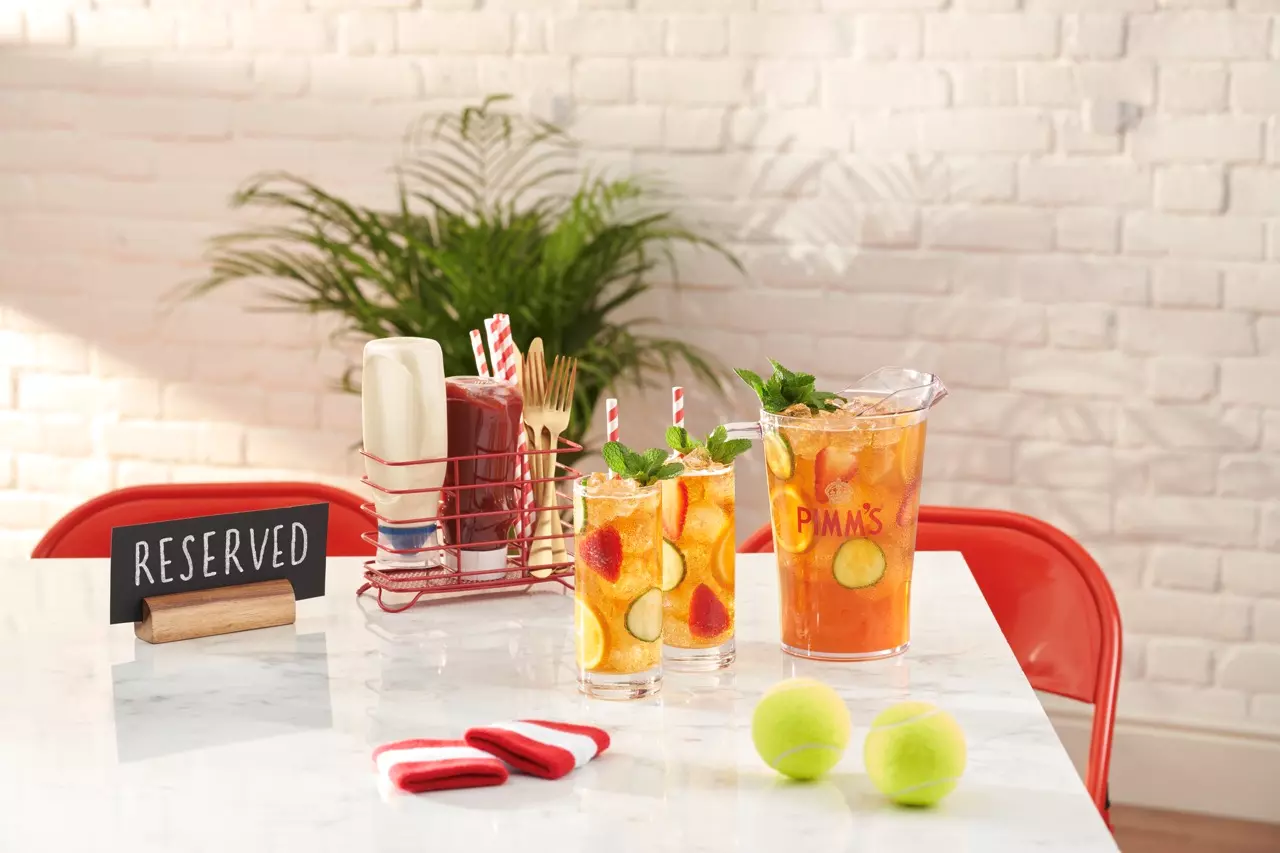 A pitcher and two glasses of Pimm’s cocktail with fruit and mint on a marble table, near tennis balls, utensils, and a reserved sign, with a plant and white brick wall in the background.