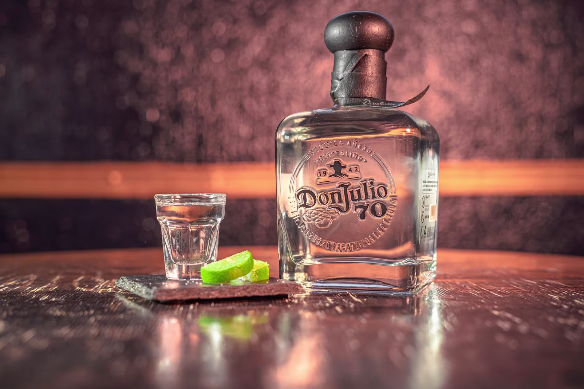 A bottle of Don Julio tequila sits on a wooden table next to a shot glass and lime wedges on a small slate tray.