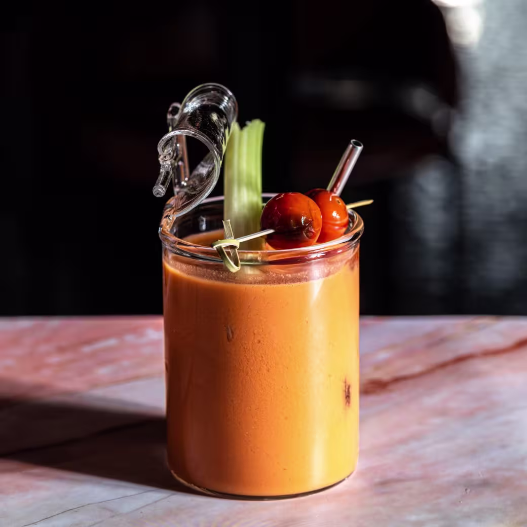 La foto muestra un vaso con un cóctel de color naranja, adornado con un tallo de apio y dos aceitunas ensartadas. La bebida está sobre una mesa con una superficie rosada, y el fondo es oscuro, lo que resalta el color vibrante de la bebida