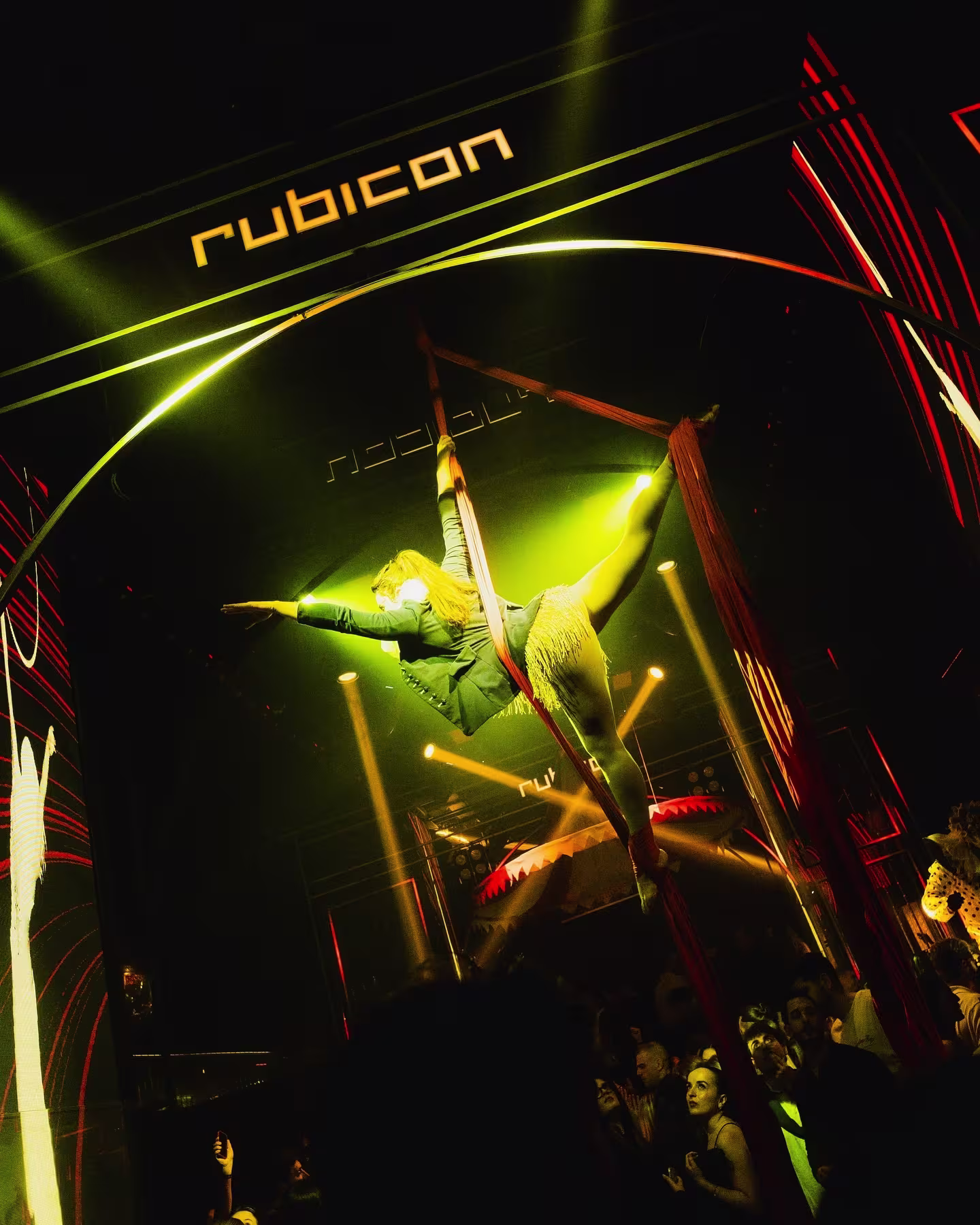 Acróbata en telas aéreas en Rubicon con luces verdes y rojas. Público disfrutando del espectáculo en una noche vibrante y llena de energía.