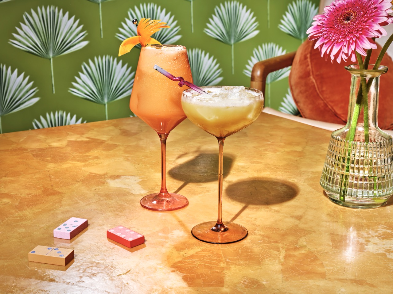 Two colorful cocktails in stemmed glasses on a marble table, with dominoes and a vase of pink flowers, against a green palm-patterned wallpaper background.