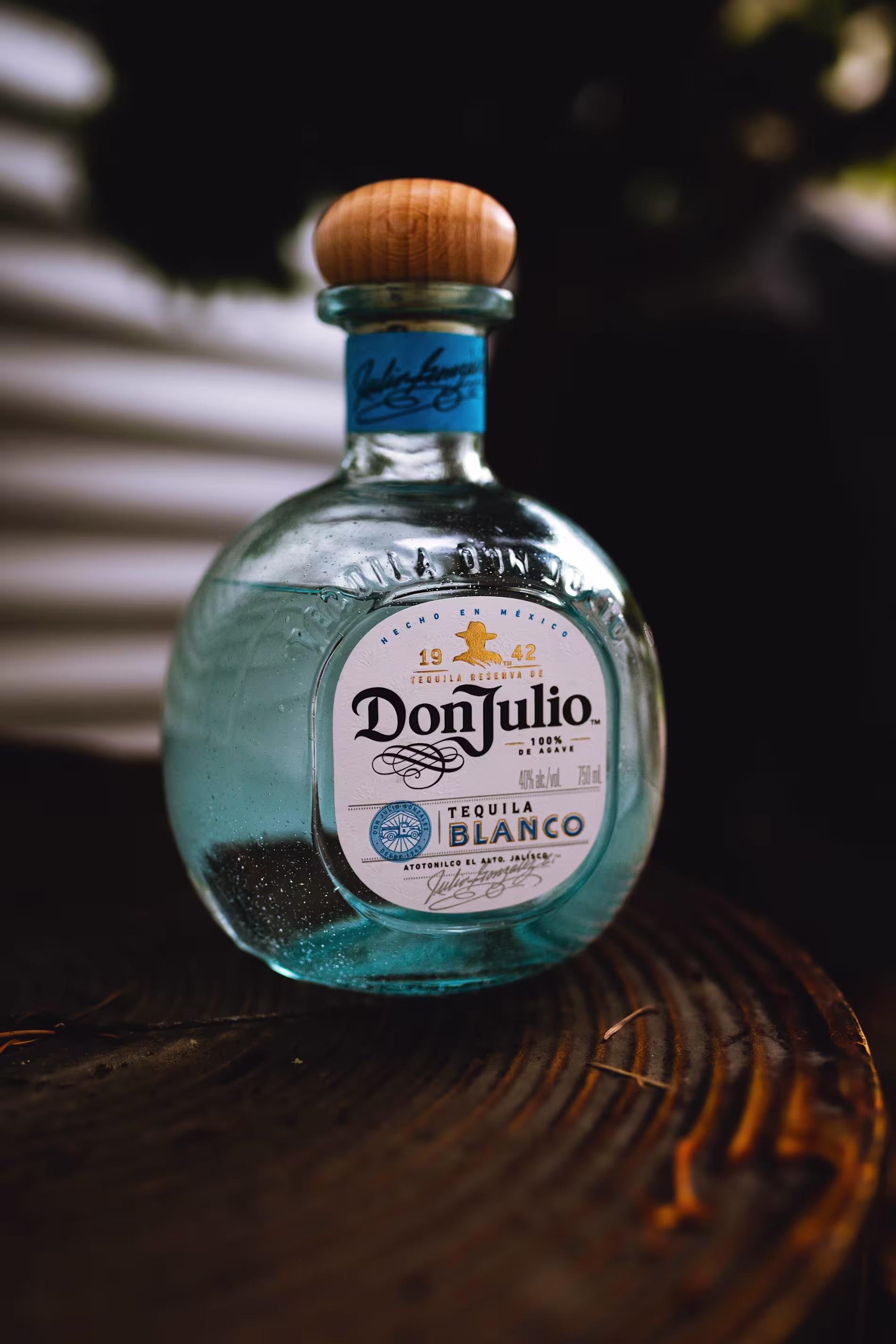 A bottle of Don Julio Tequila Blanco with a wooden cap sits on a dark wooden surface.