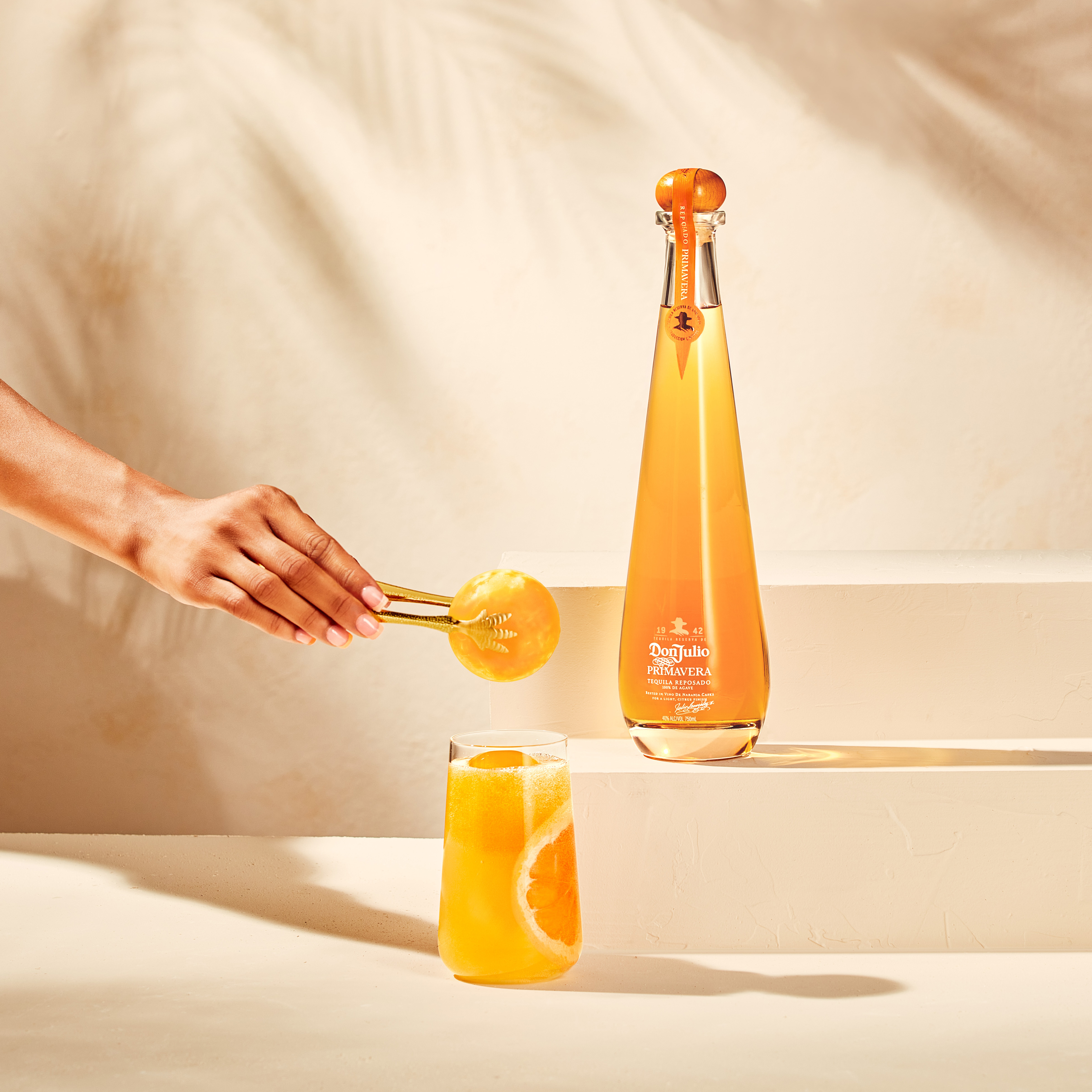 A hand garnishes an orange cocktail with a slice of orange next to a tall bottle of Don Julio Primavera tequila on a light-colored surface.
