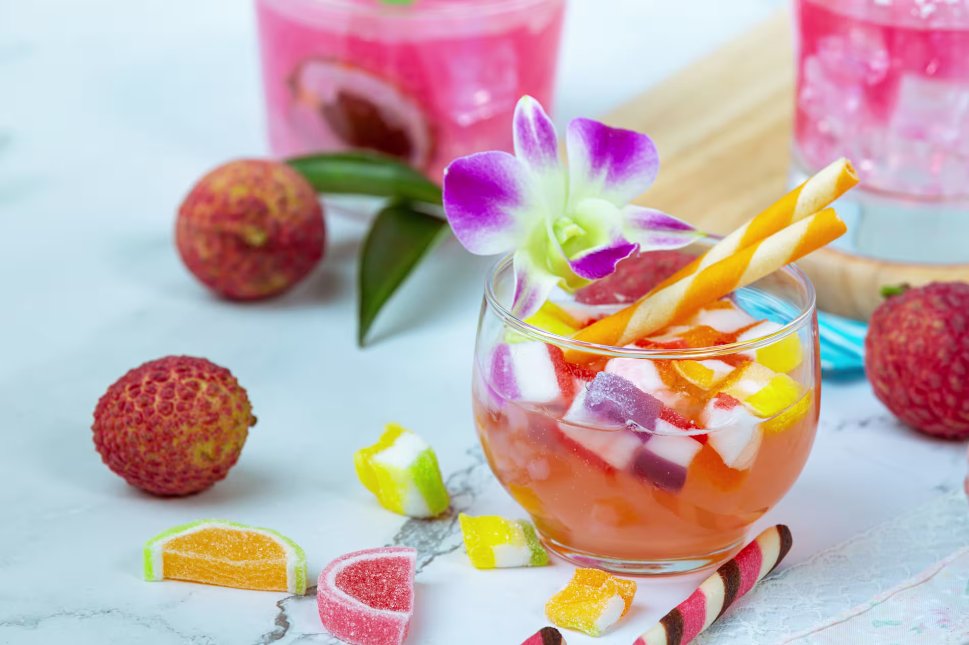 A glass of colorful fruit punch with ice, jelly candies, a striped straw, and a purple orchid garnish, surrounded by lychees and assorted gummy candies on a marble surface.