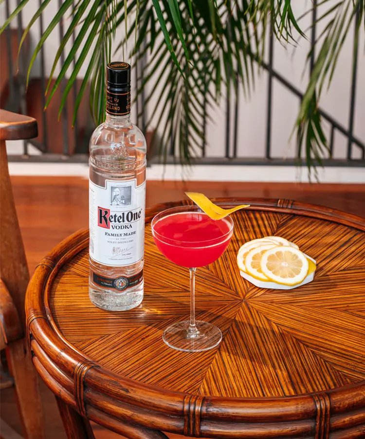 A bottle of Ketel One vodka, a red cocktail with a lemon twist, and lemon slices on a wooden table with a leafy plant in the background.