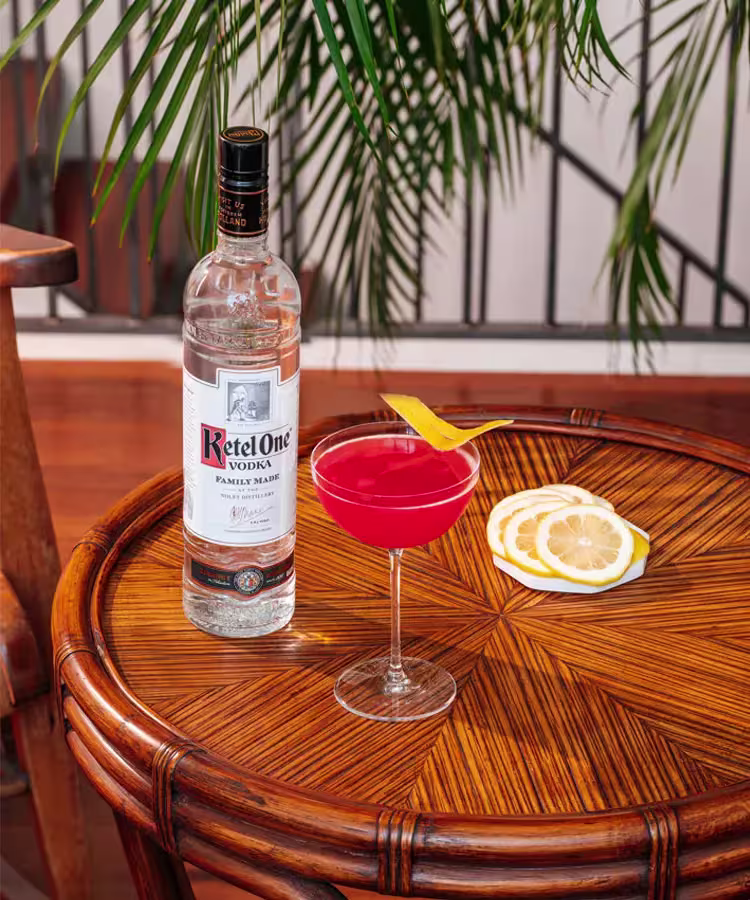 A bottle of Ketel One vodka, a red cocktail with a lemon twist, and lemon slices on a wooden table with a leafy plant in the background.