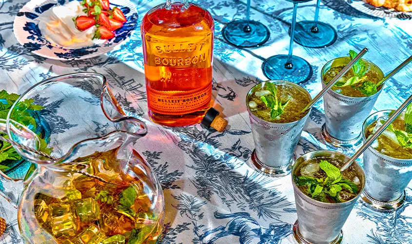 A table set with a bottle of Bulleit Bourbon, a glass pitcher of a drink with ice, four mint juleps in metal cups, and a plate with strawberries and cream on a blue-patterned tablecloth.