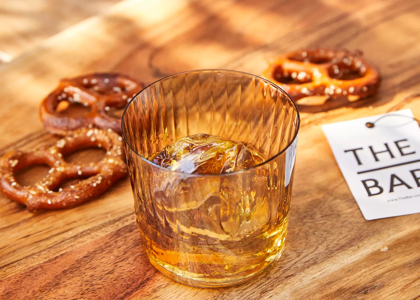 A glass of whiskey with a large ice cube is on a wooden surface next to three pretzels and a card labeled THE BAR.