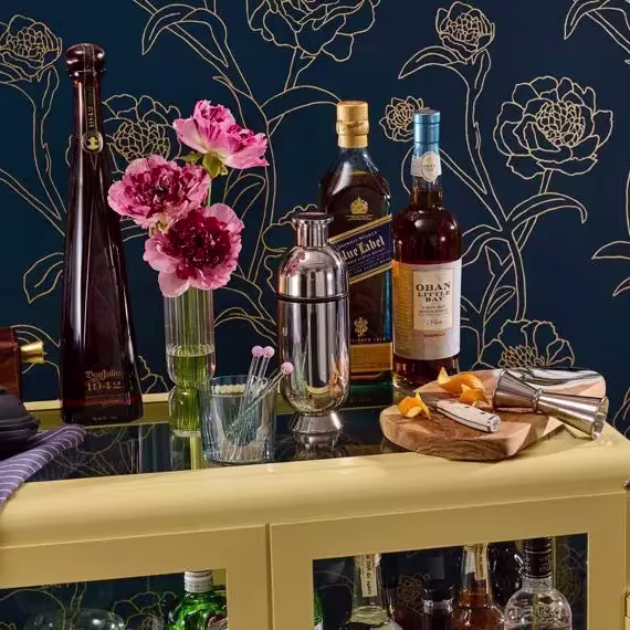 A bar cart with liquor bottles, a shaker, a vase of pink flowers, cocktail picks, a cutting board with orange peels, and a jigger against a floral-patterned wall.