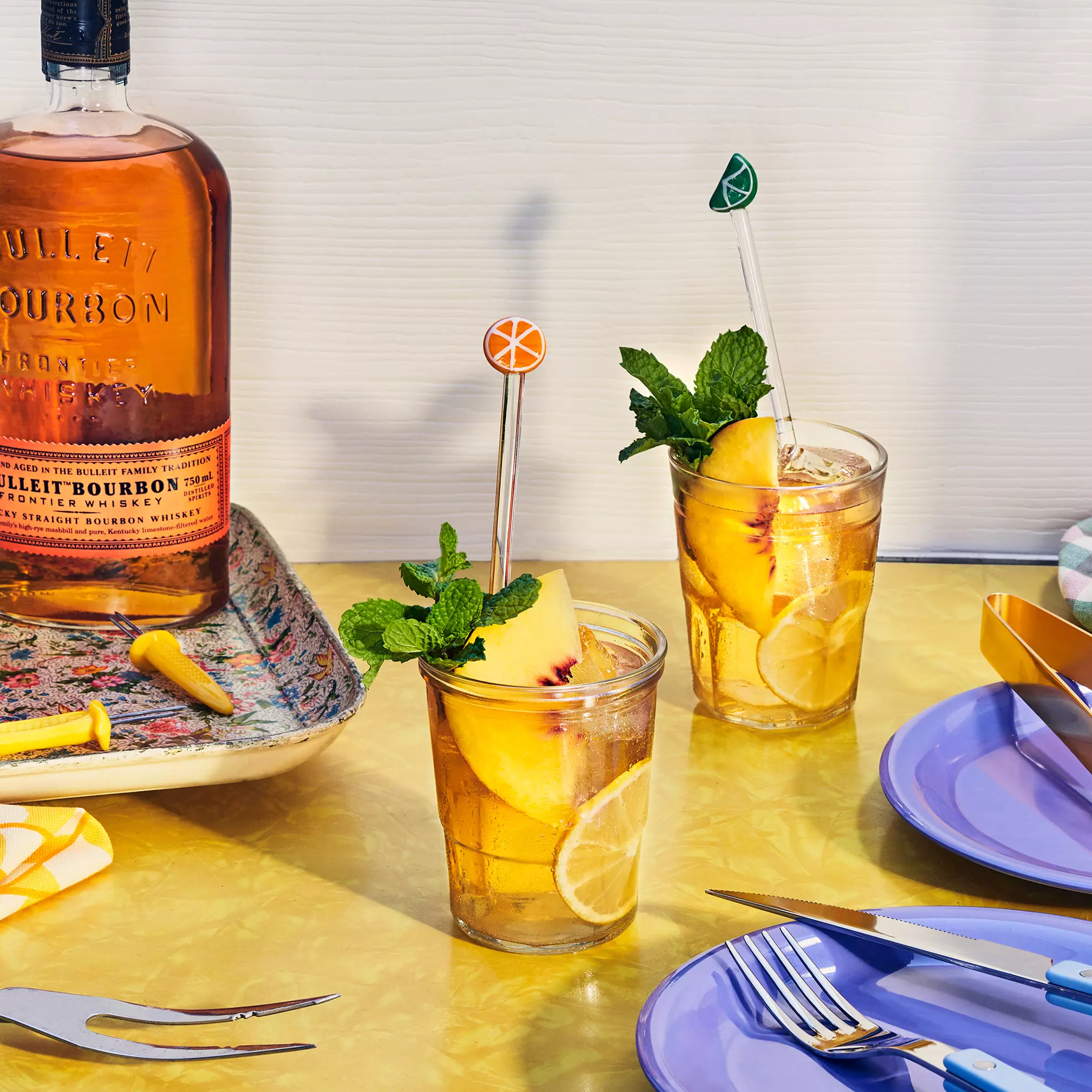 Two glasses of bourbon cocktails with lemon slices, mint, and fruit garnishes sit on a yellow table next to a bottle of Bulleit Bourbon and a platter with lemon twists.