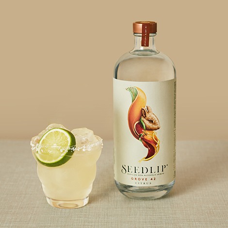 A margarita in a rocks glass with a salt rim and lime wheel garnish. Sitting next to a bottle of Seedlip Grove.