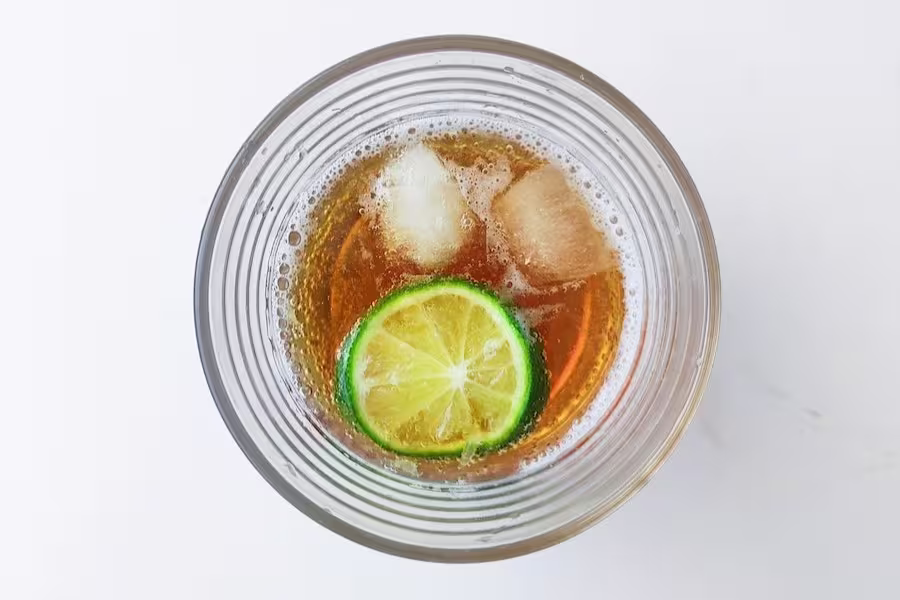 A glass of brown beverage with ice cubes and a slice of lime, viewed from above, on a white background.