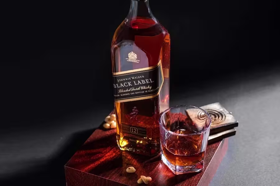 A bottle of Johnnie Walker Black Label whisky and a glass of whisky sit on a wooden surface with a few scattered nuts against a dark background.