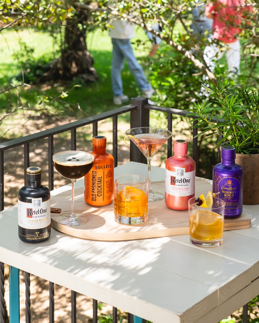 A tray with five cocktail glasses and five bottles of spirits, including Ketel One and Bulleit, on an outdoor table next to a railing and potted plant.