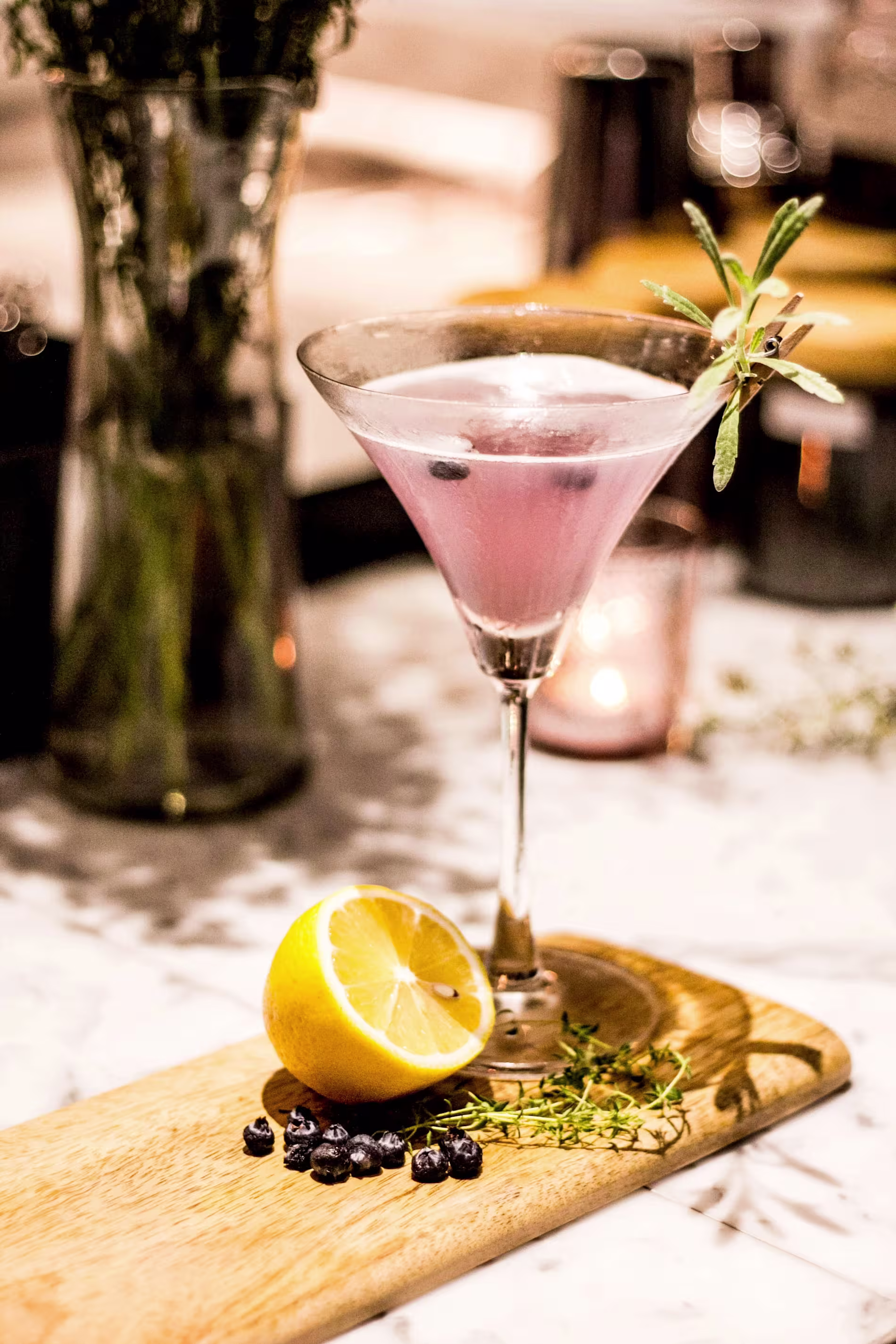 A pink cocktail in a martini glass garnished with herbs, beside a halved lemon, blueberries, and sprigs of thyme on a wooden board.