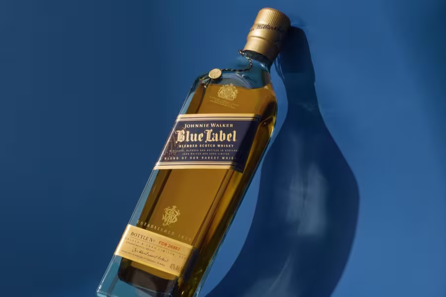 A bottle of Johnnie Walker Blue Label blended Scotch whisky lies on its side against a solid blue background, casting a shadow.
