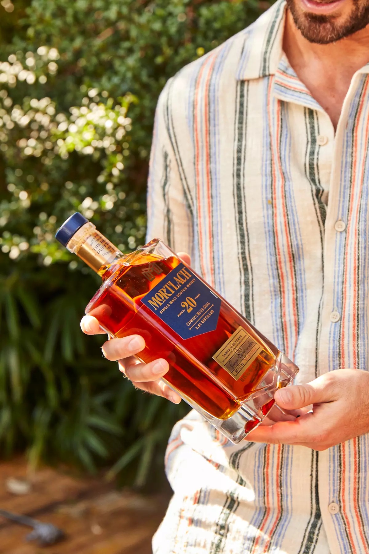 A person in a striped shirt holds a bottle of Mortlach 20-year-old single malt Scotch whisky outdoors.