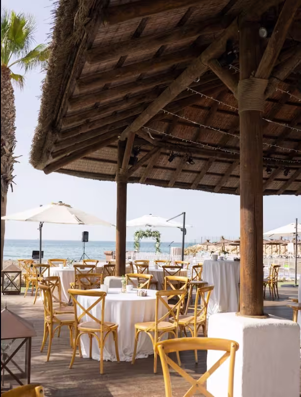 La imagen muestra el restaurante Candado Beach junto a la playa. Hay mesas de madera con sillas, manteles blancos y grandes sombrillas que proporcionan sombra. El suelo parece ser arenoso y el océano es visible en el fondo
