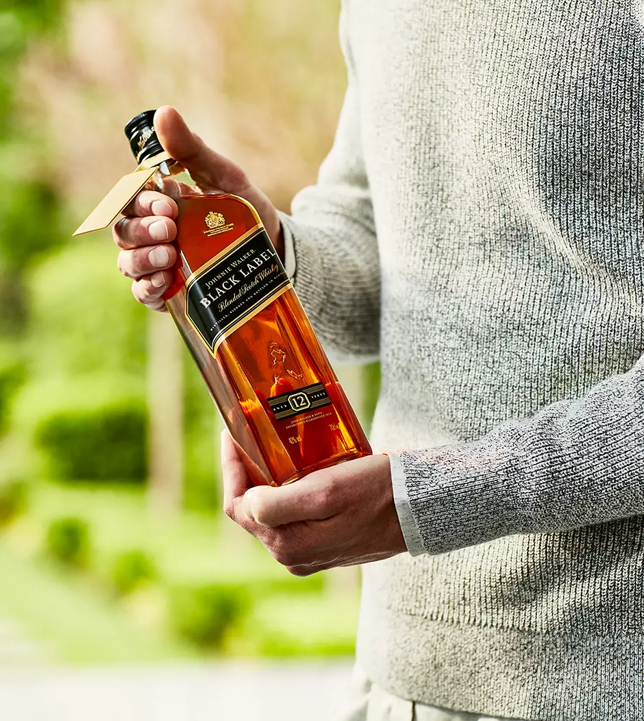 A person in a gray sweater holds a bottle of Johnnie Walker Black Label 12-year-old blended Scotch whisky outdoors.