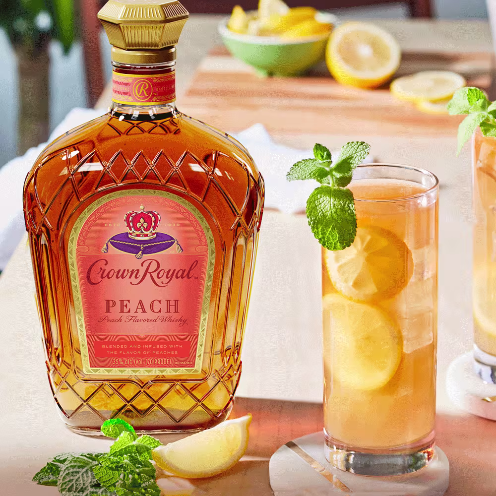 A bottle of Crown Royal Peach whiskey beside a glass of iced tea garnished with lemon slices and mint, with lemon wedges and a cutting board in the background.