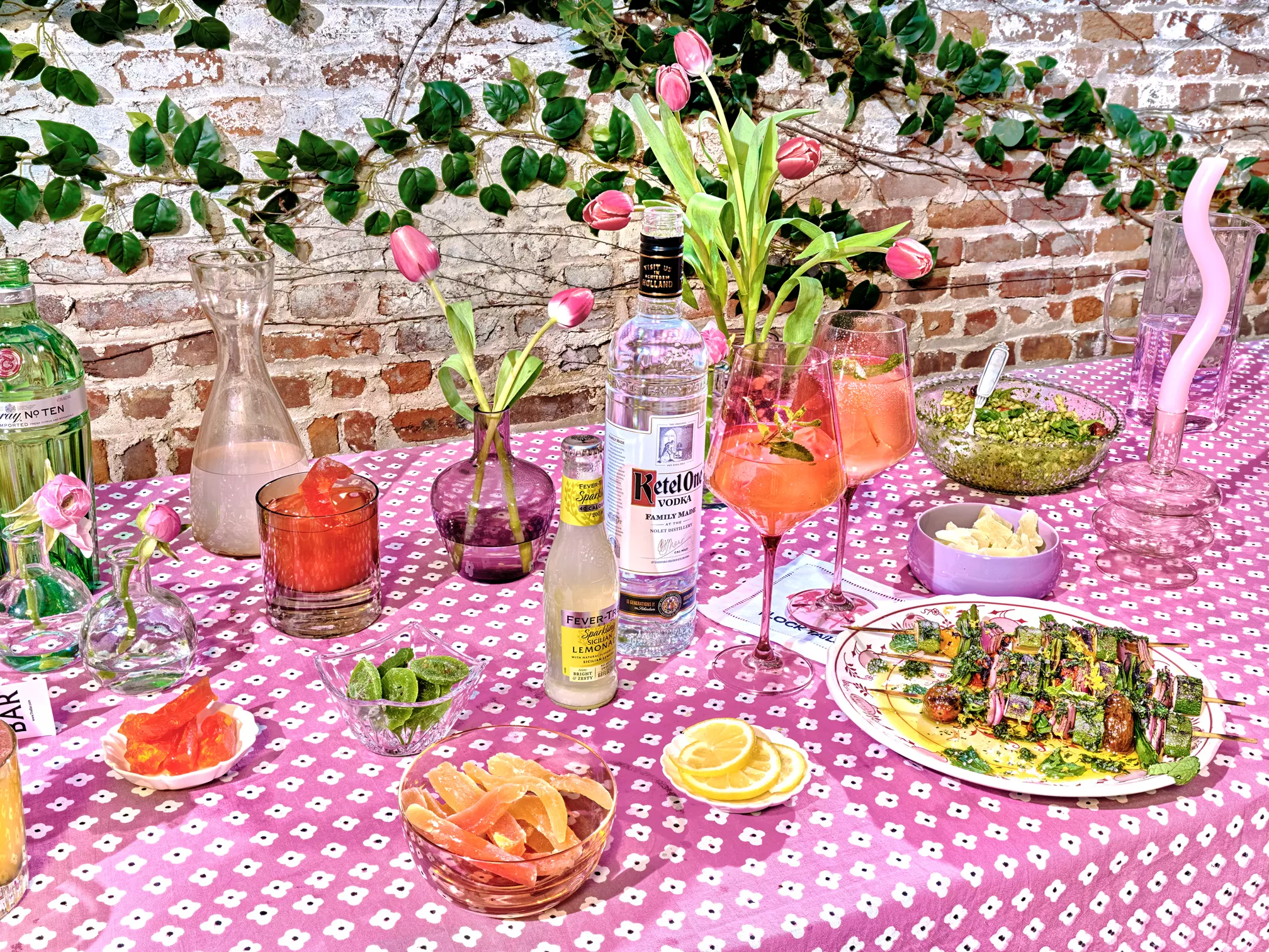 A table with pink-patterned cloth displays cocktails, bottled drinks, sliced citrus, vases with flowers, snacks, and a plate of food against a brick wall with greenery.