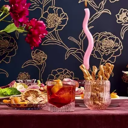 A table set with drinks, breadsticks in a glass, assorted dried fruits, and flowers, with a pink twisted candle and floral wallpaper in the background.