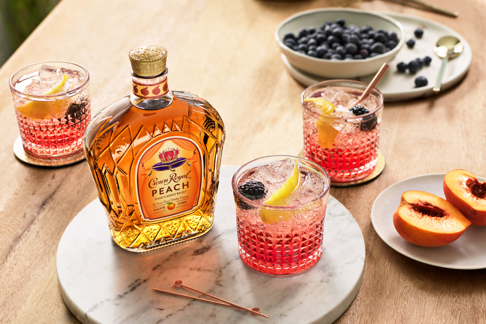 A bottle of Crown Royal Peach whiskey sits on a marble tray with three peach cocktails, fresh peaches, and a bowl of blueberries on a wooden table.