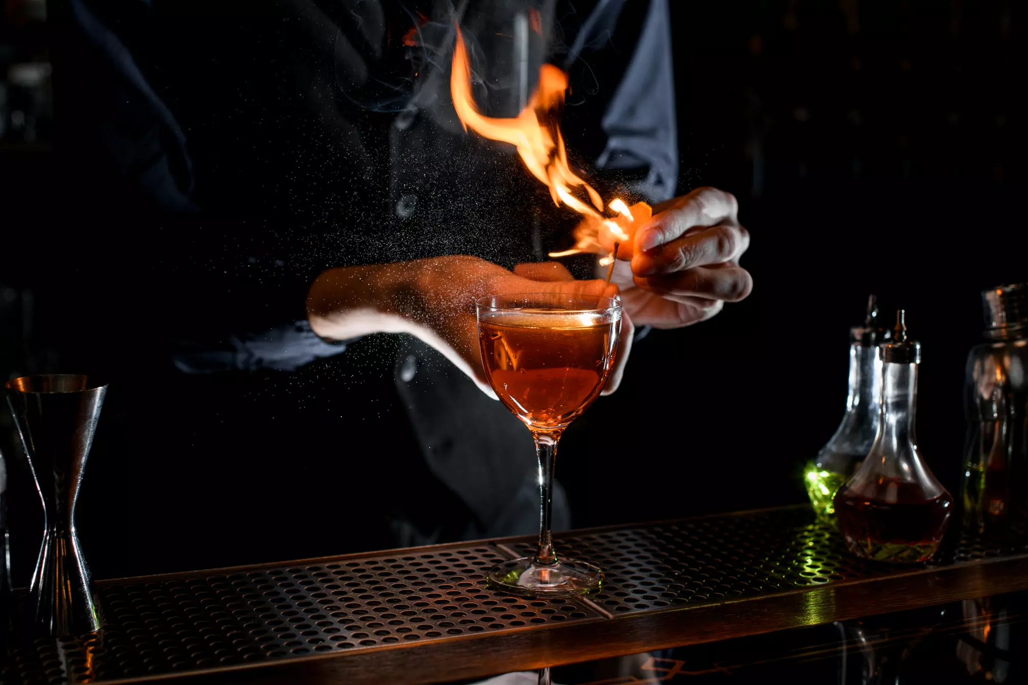 A bartender ignites a flame above a cocktail glass, creating a dramatic presentation at a dimly lit bar counter.