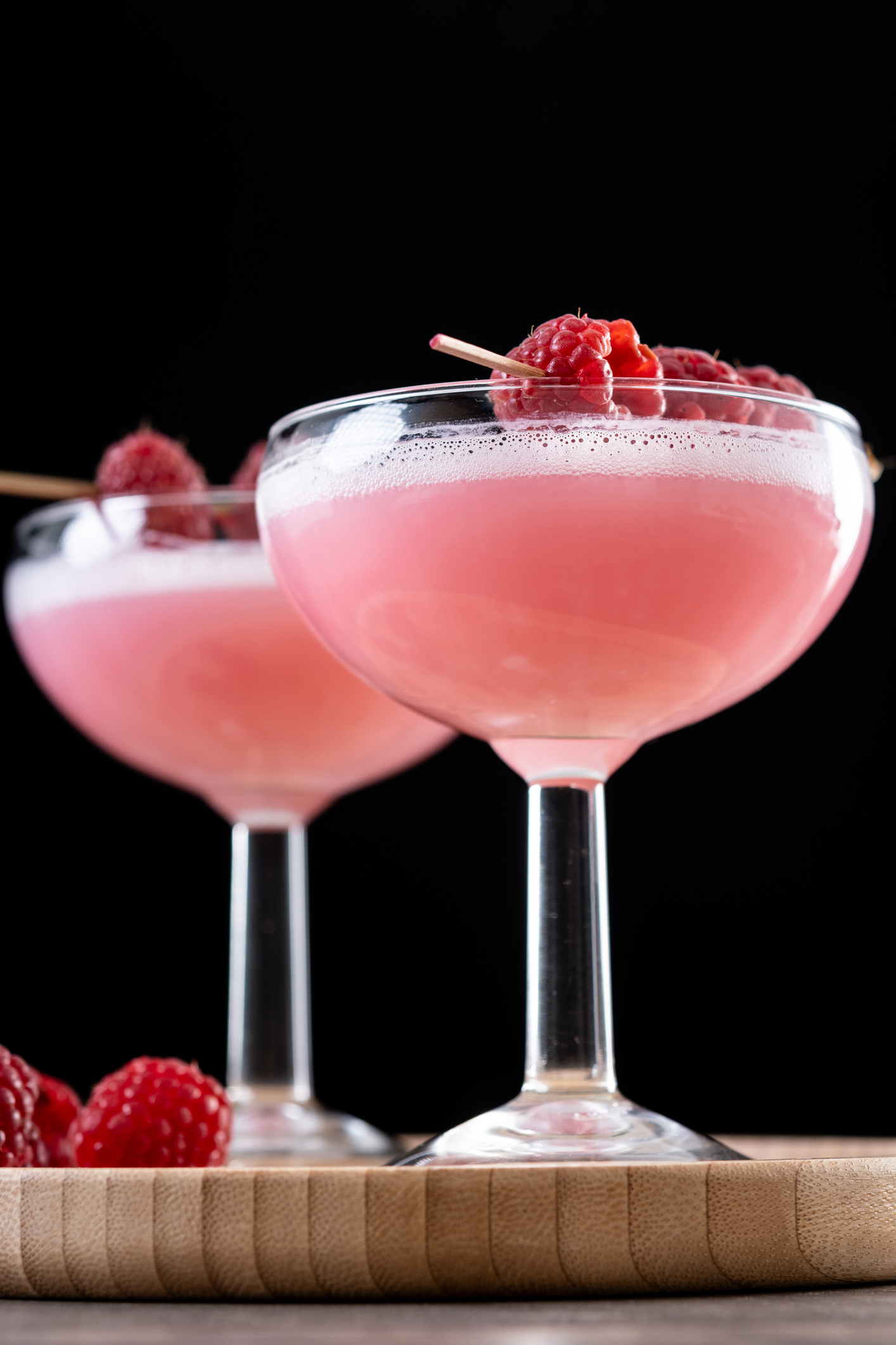 Two coupe glasses filled with pink cocktails, garnished with fresh raspberries on a skewer, are set against a black background.