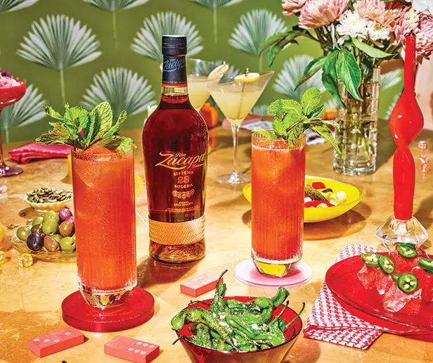 A bottle of Zacapa rum on a table with two tall red cocktails, snacks, flowers, olives, and a martini, set against a patterned green background.