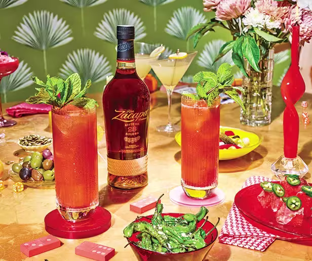 A bottle of Zacapa rum on a table with two tall red cocktails, snacks, flowers, olives, and a martini, set against a patterned green background.