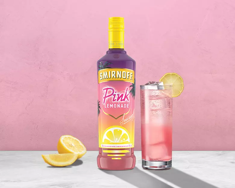 A bottle of Smirnoff Pink Lemonade, a glass with pink drink and lemon slice, and lemon wedges on a gray surface against a pink background.