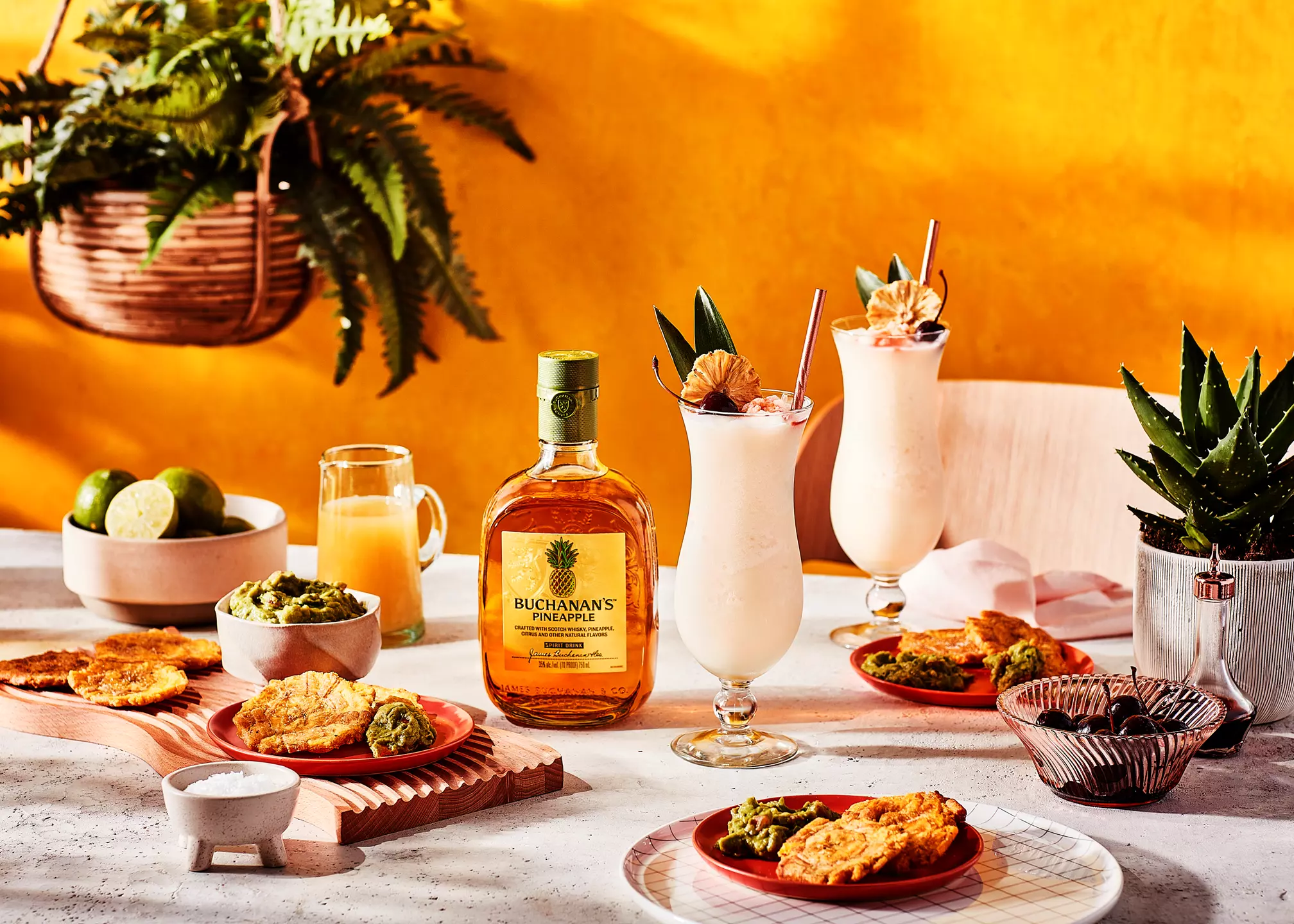 A table with Buchanan’s Pineapple bottle, two creamy cocktails, guacamole, fried snacks, limes, juice, and decorative plants against a bright orange background.