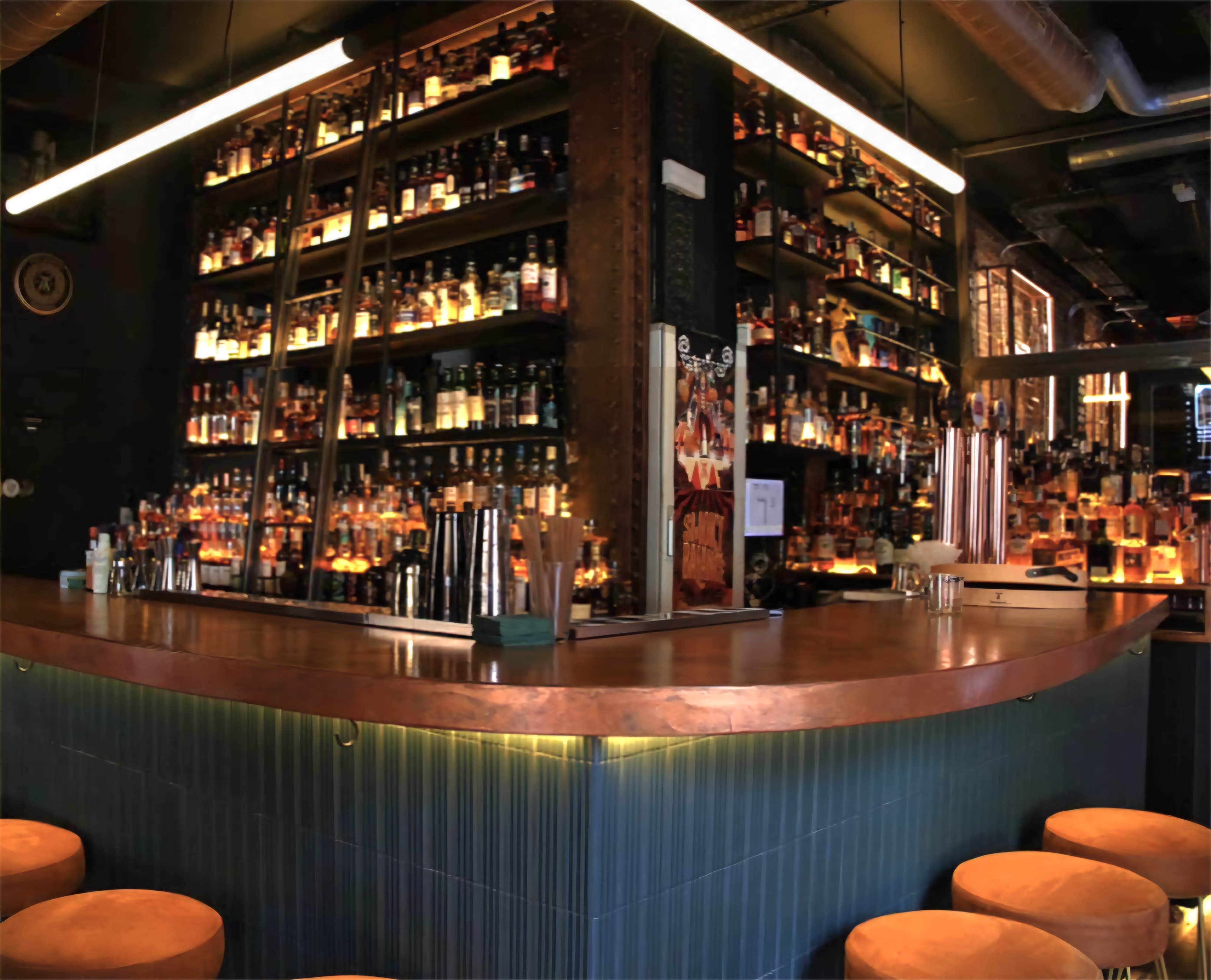 La imagen muestra el bar Holy Drop, concurrido con varias personas reunidas alrededor del mostrador. Detrás del mostrador, hay varias botellas de alcohol exhibidas en estantes y un gran logo en la pared.