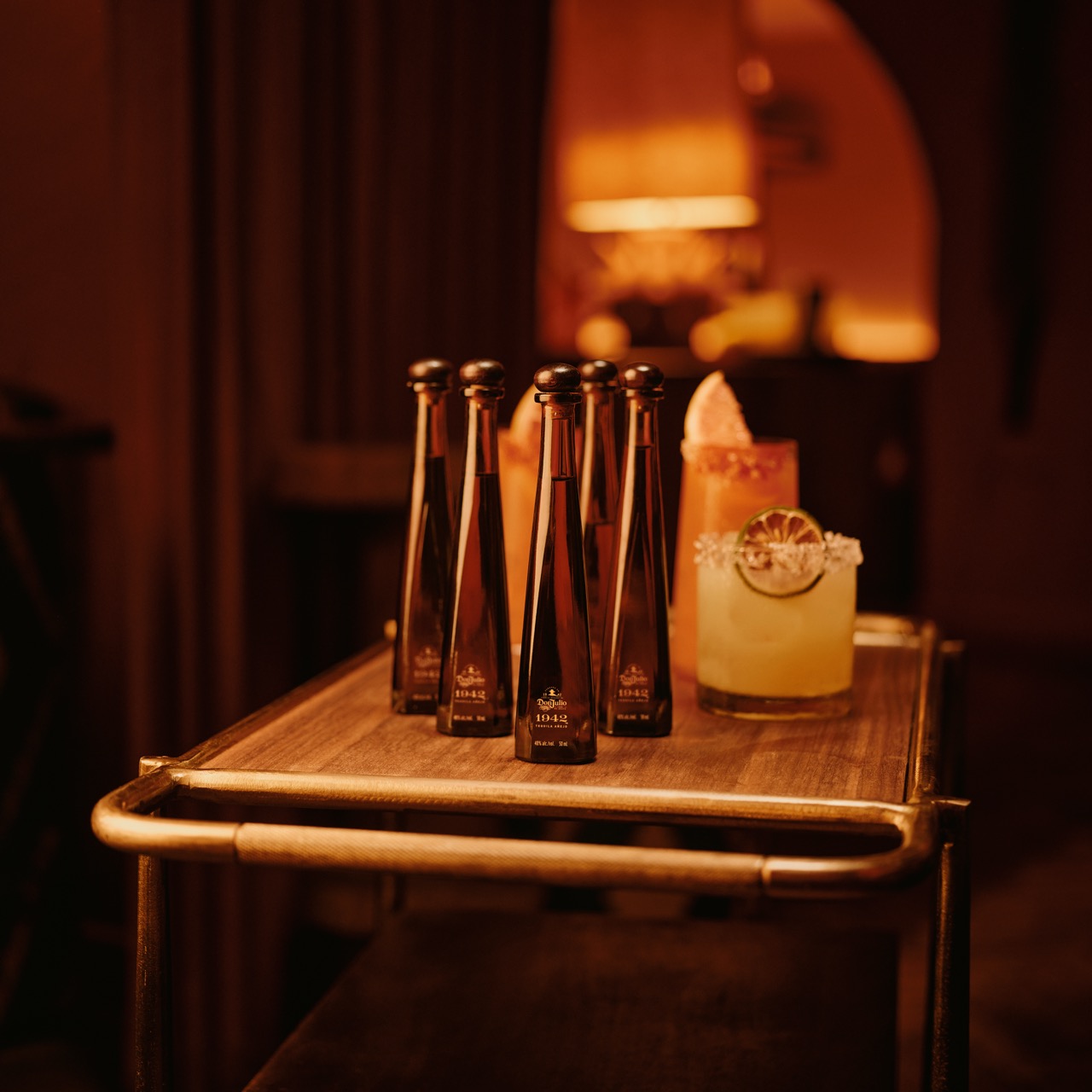 Six tall, narrow bottles and two cocktails with garnishes are arranged on a wooden cart in a warmly lit room.