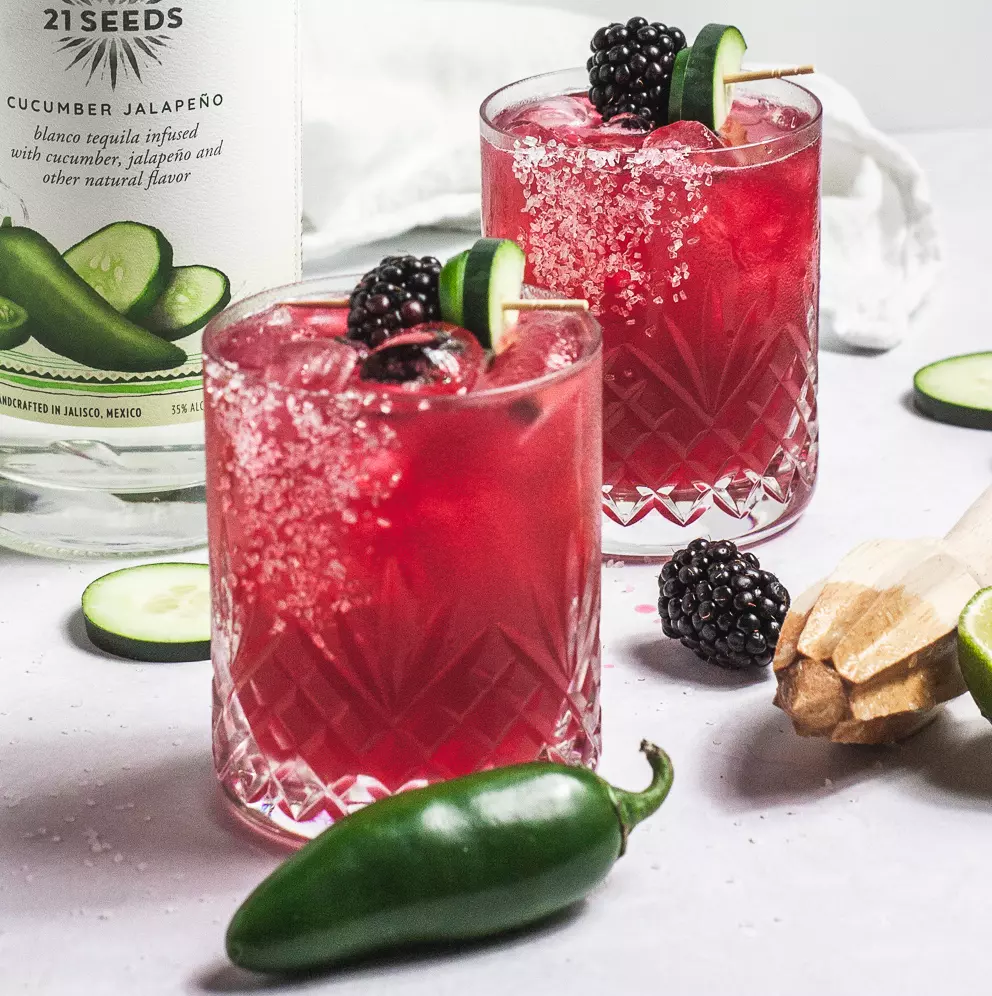 Two glasses of a bright pink cocktail garnished with blackberries and cucumber slices, with a jalapeño, cucumber slices, and a tequila bottle nearby.
