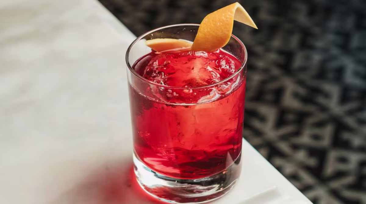A glass filled with a red cocktail over ice, garnished with an orange twist, sits on a light-colored surface.
