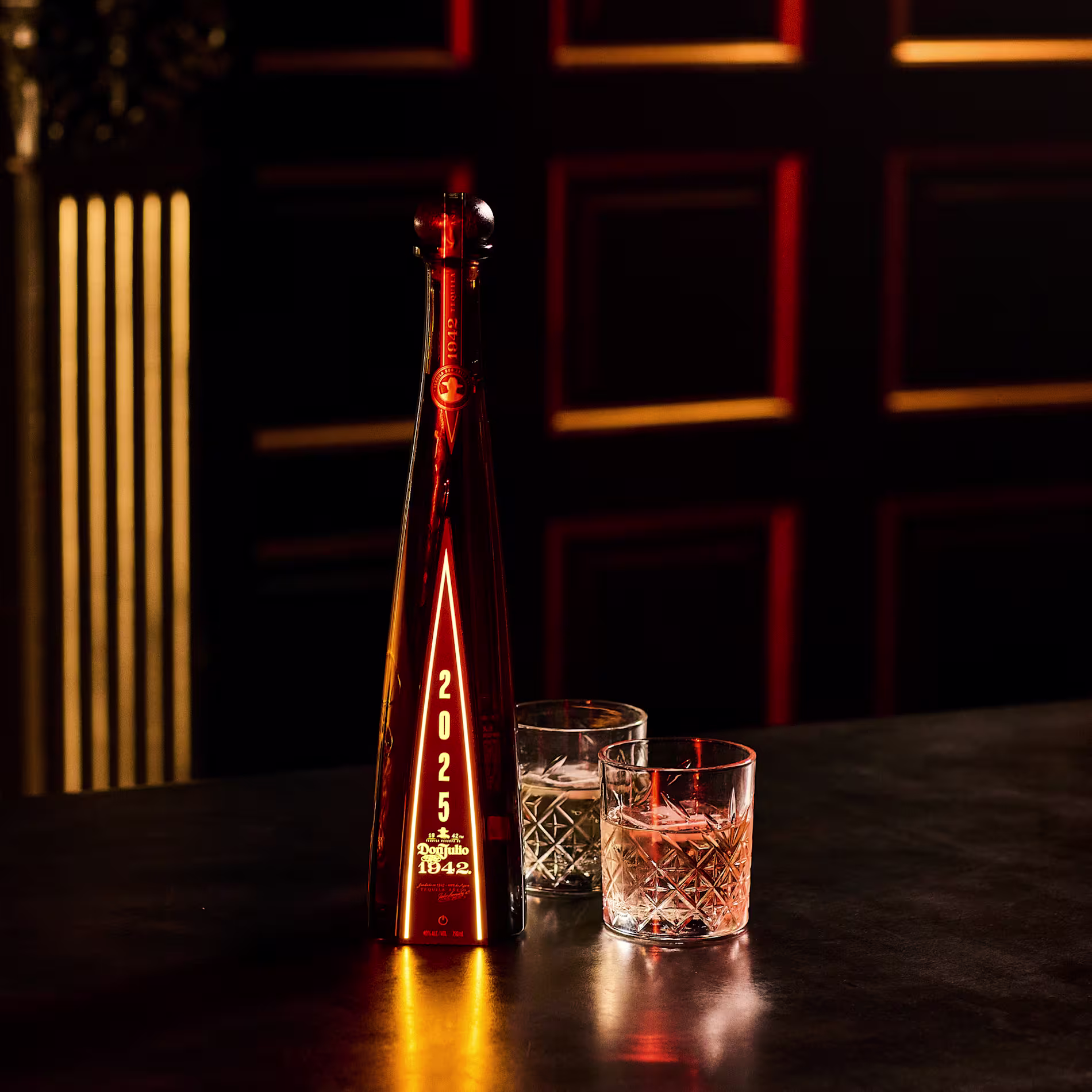 A tall, amber bottle labeled 2025 stands on a dark table beside two crystal glasses, one filled with clear liquid and the other with amber liquid, against a dark, ornate background.