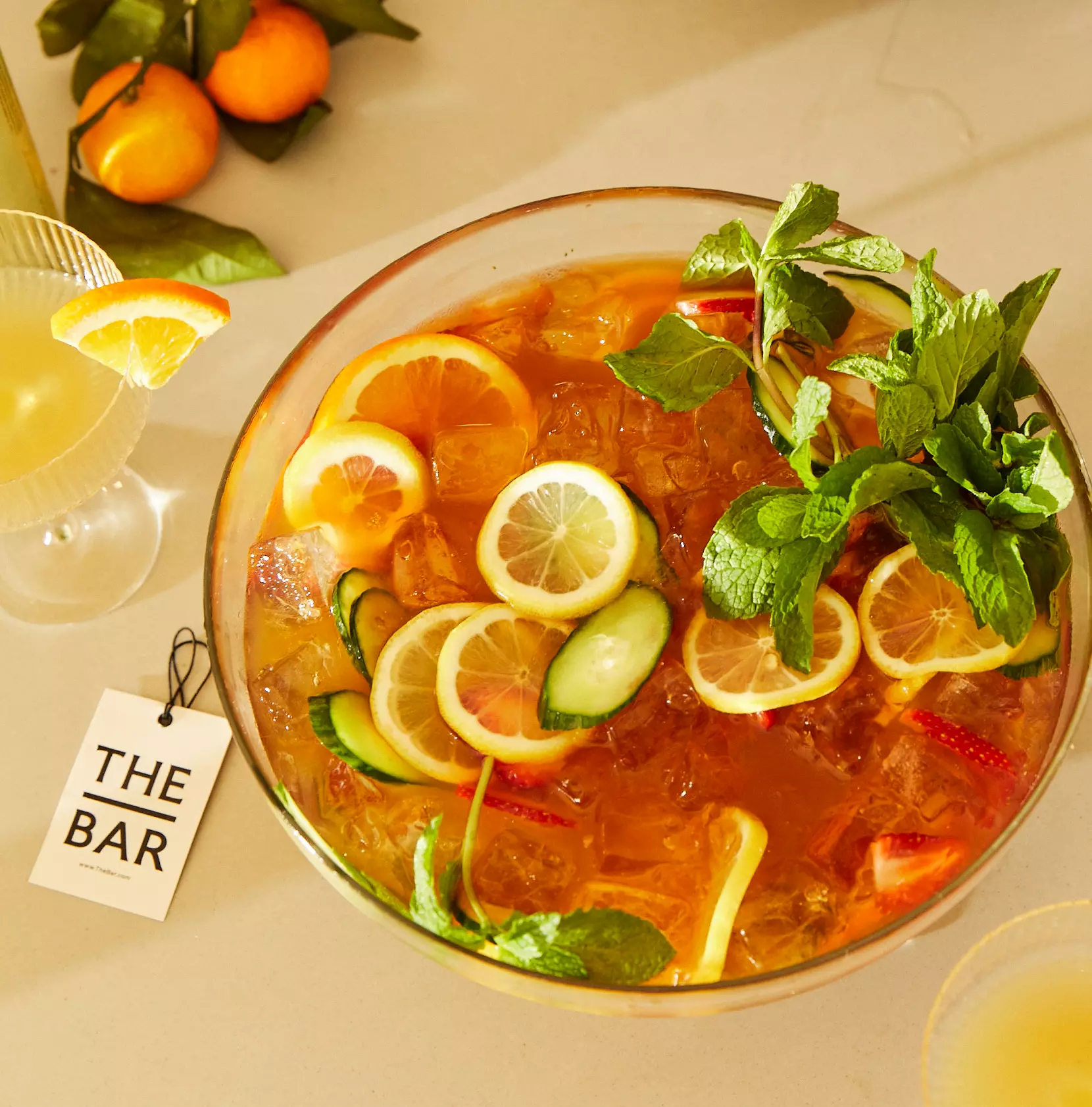 A bowl of iced punch garnished with lemon, cucumber, strawberries, and mint sits on a table next to a cocktail and a tag labeled THE BAR.
