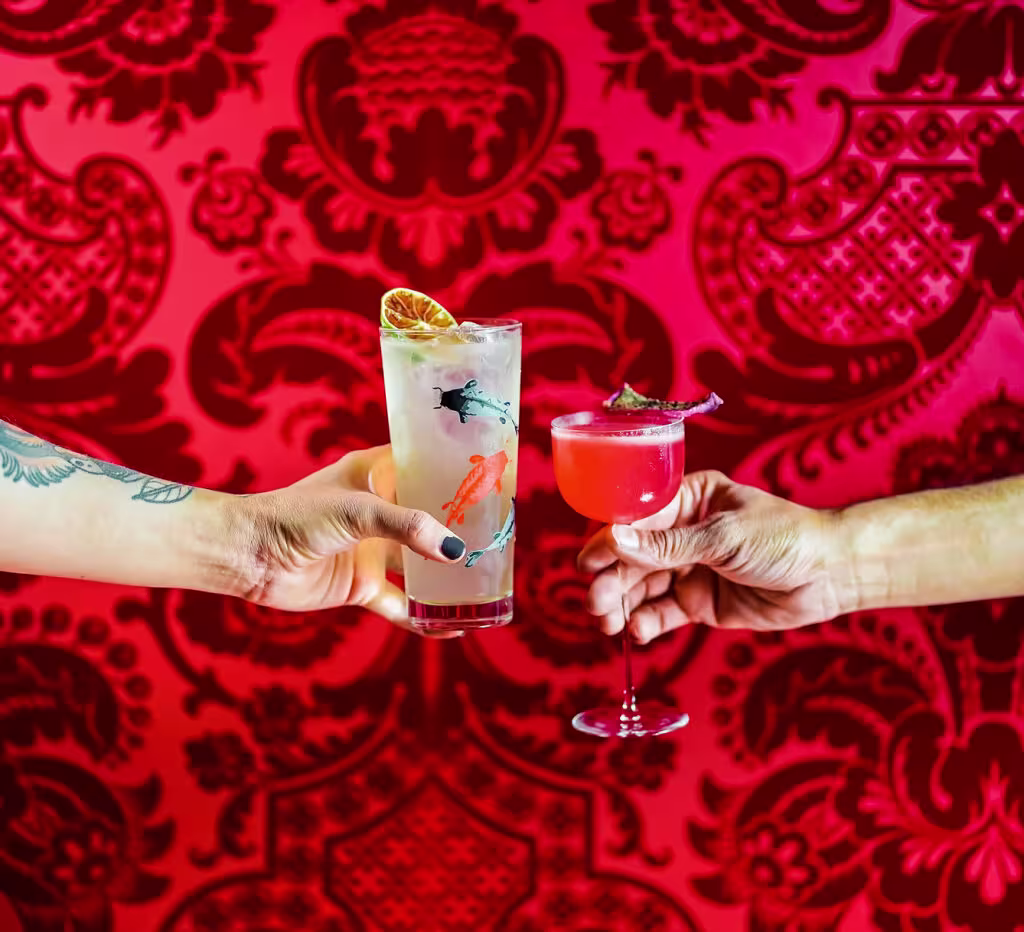 Dos manos brindan con cócteles coloridos frente a un vibrante fondo rojo con estampado floral, evocando un ambiente de celebración y estilo.