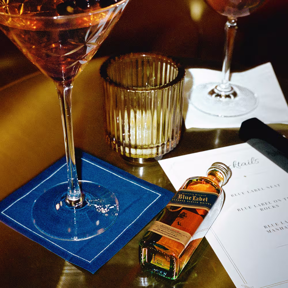 A small bottle of Johnnie Walker Blue Label, two cocktail glasses, a candle, and a drink menu on a gold table.