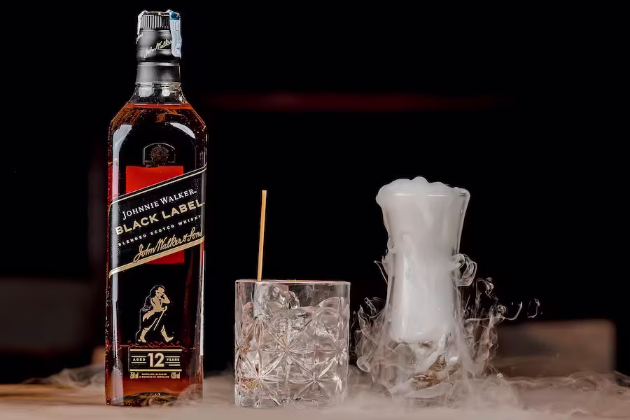 A bottle of Johnnie Walker Black Label whisky stands next to a glass with whisky and ice, and a foggy cocktail in a tall glass with vapor rising.
