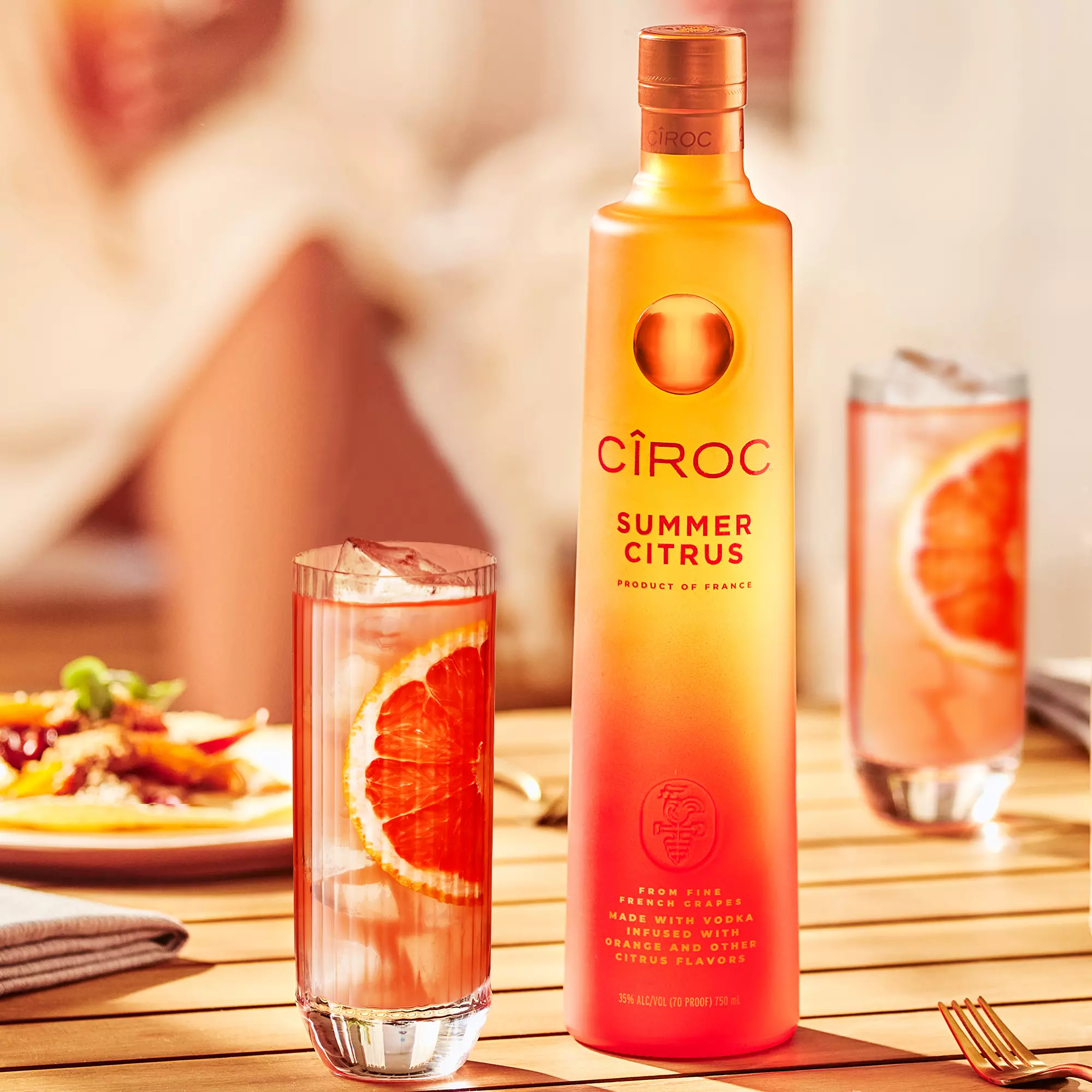 A Ciroc Summer Citrus vodka bottle stands on a wooden table next to a tall glass with ice and a slice of orange; blurred food and drink are in the background.