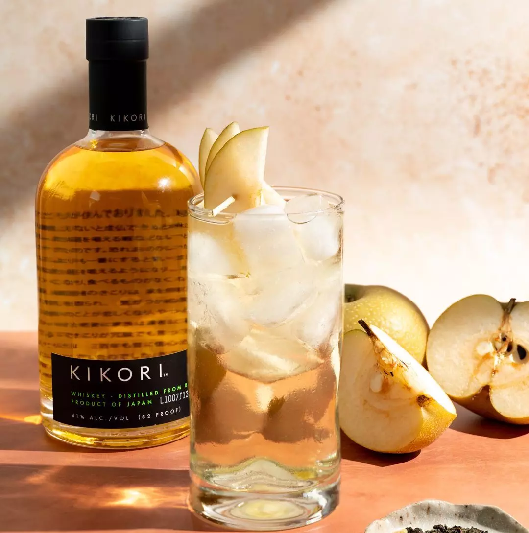 A bottle of Kikori whiskey stands beside a glass of whiskey with ice and pear slices; fresh and sliced pears are on the table.