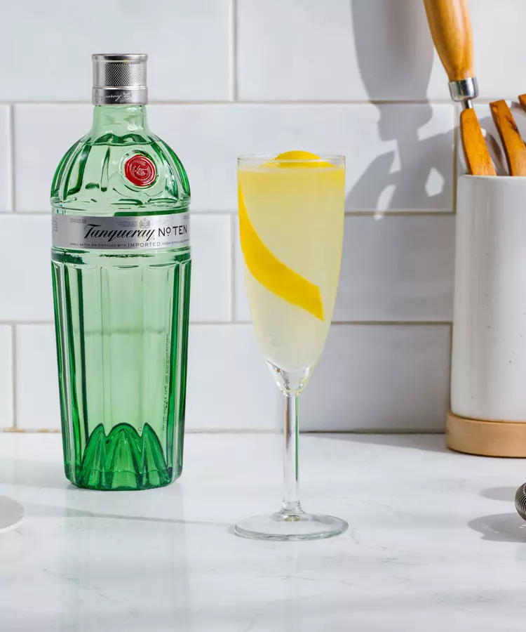A bottle of Tanqueray No. Ten gin stands next to a champagne flute filled with a yellow cocktail garnished with a lemon twist, set on a white kitchen counter.