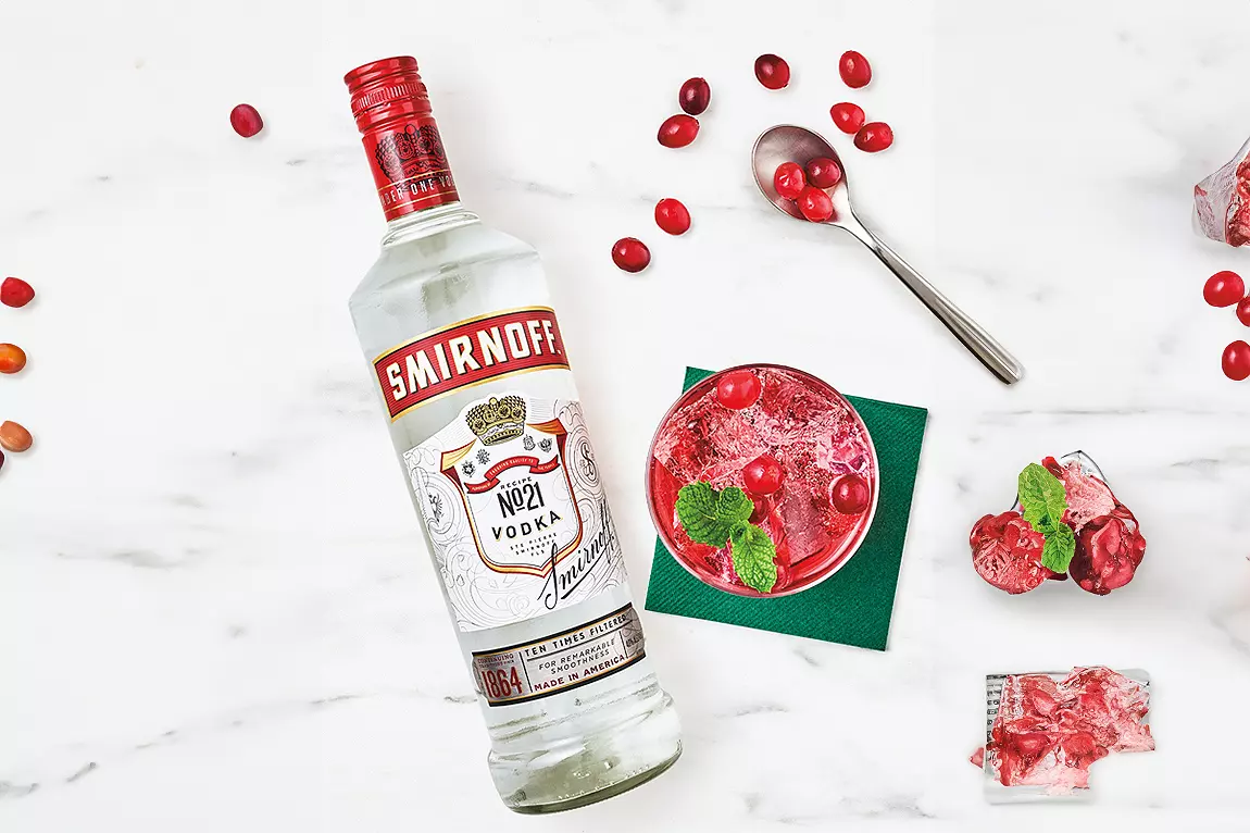 A bottle of Smirnoff vodka lies next to a red cocktail with cranberries and mint, a spoon with cranberries, ice cubes, and a green napkin on a white surface.