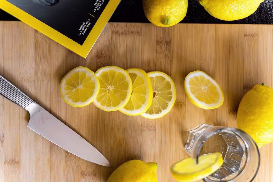 Sliced lemons on a wooden cutting board beside a knife, whole lemons, a glass with a lemon slice, and a yellow-edged book.
