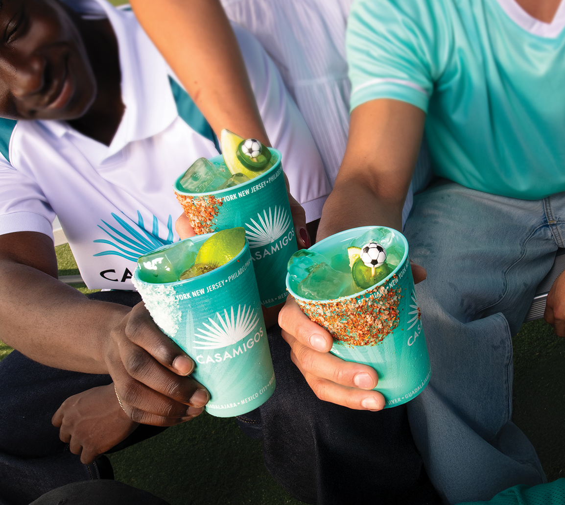 Three people toast with turquoise Casamigos cups filled with blue cocktails, lime wedges, colorful rims, and small soccer ball decorations, all wearing teal or white shirts.