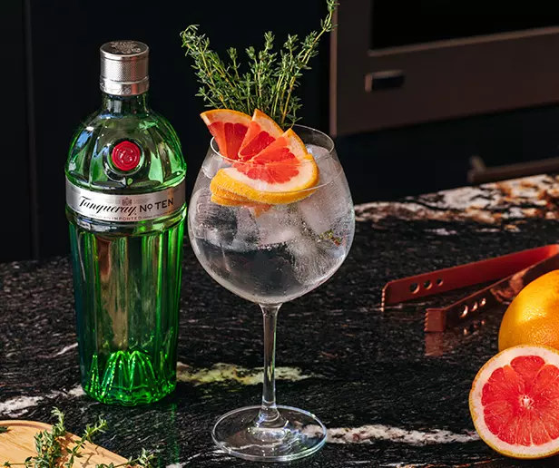 A bottle of Tanqueray No. Ten gin next to a cocktail glass filled with ice, garnished with grapefruit slices and thyme, on a dark marble countertop with grapefruit halves nearby.
